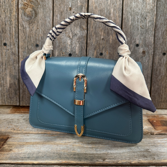 A blue leather handbag with gold hardware and a white and blue striped scarf tied around the handle, placed on a wooden surface.