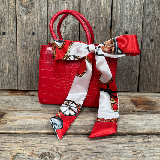 A red handbag with a white and red patterned scarf tied around it, placed on a wooden surface.