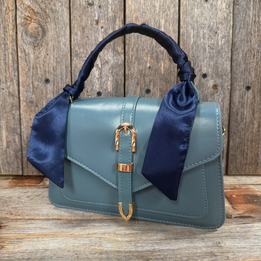 A blue satin-trimmed handbag with a gold-colored buckle and handle is displayed against a wooden background.