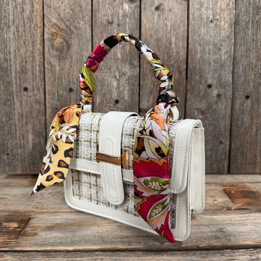 A white handbag with a colorful patterned scarf tied around the handle, placed on a wooden surface.
