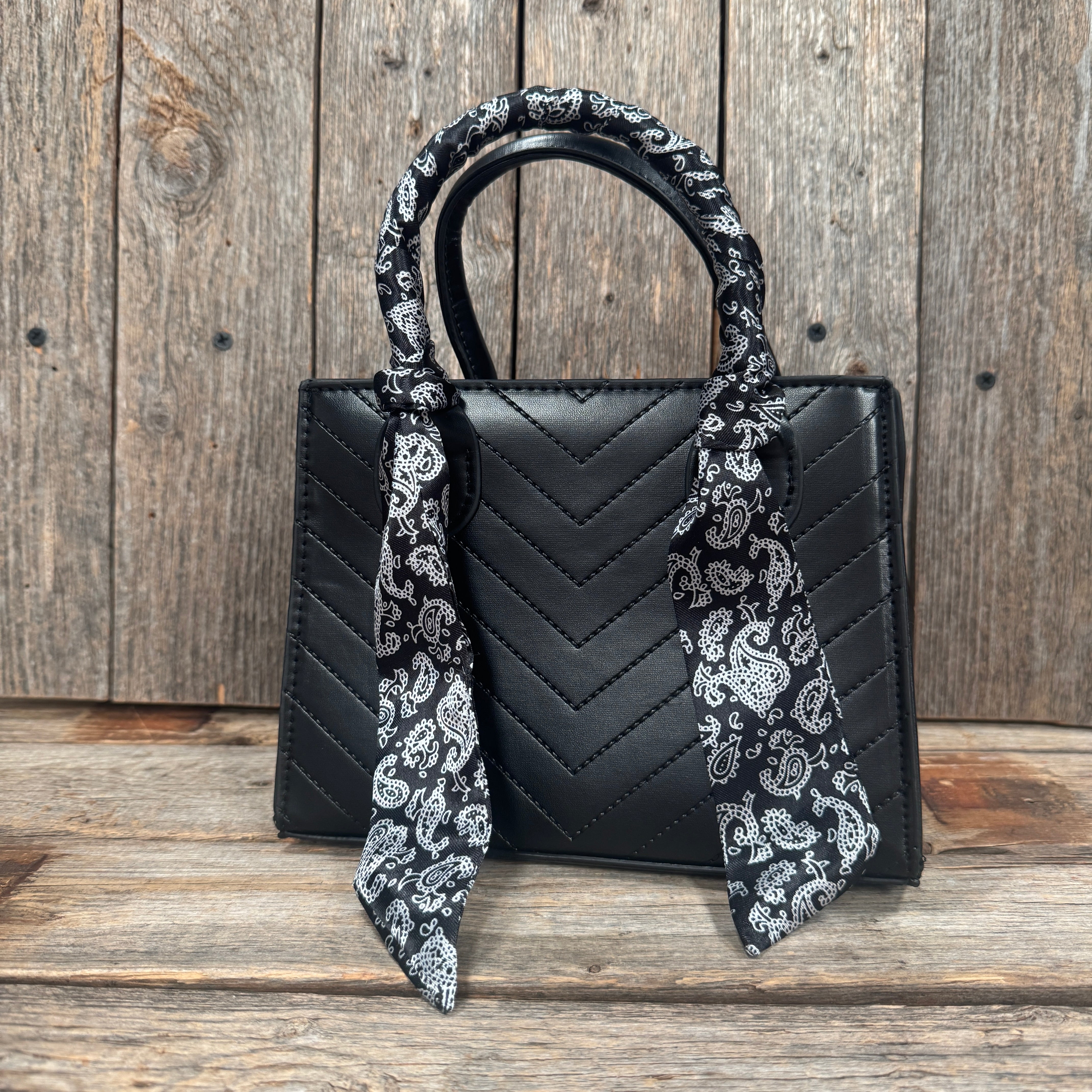A black handbag with a black and white patterned scarf tied around the handle, placed on a wooden surface.