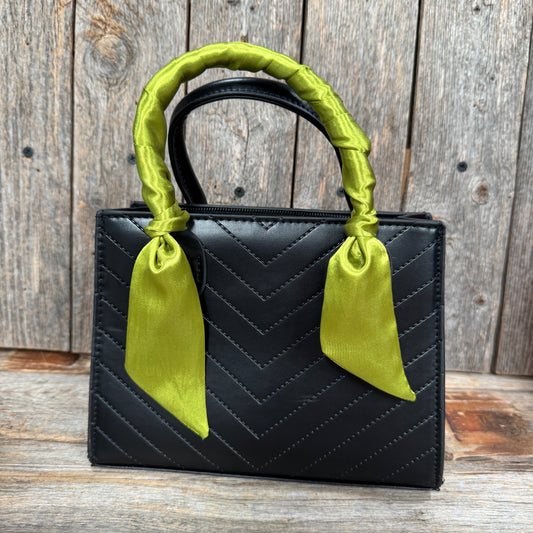 A black quilted handbag with a green ribbon tied around the handle, placed against a wooden background.