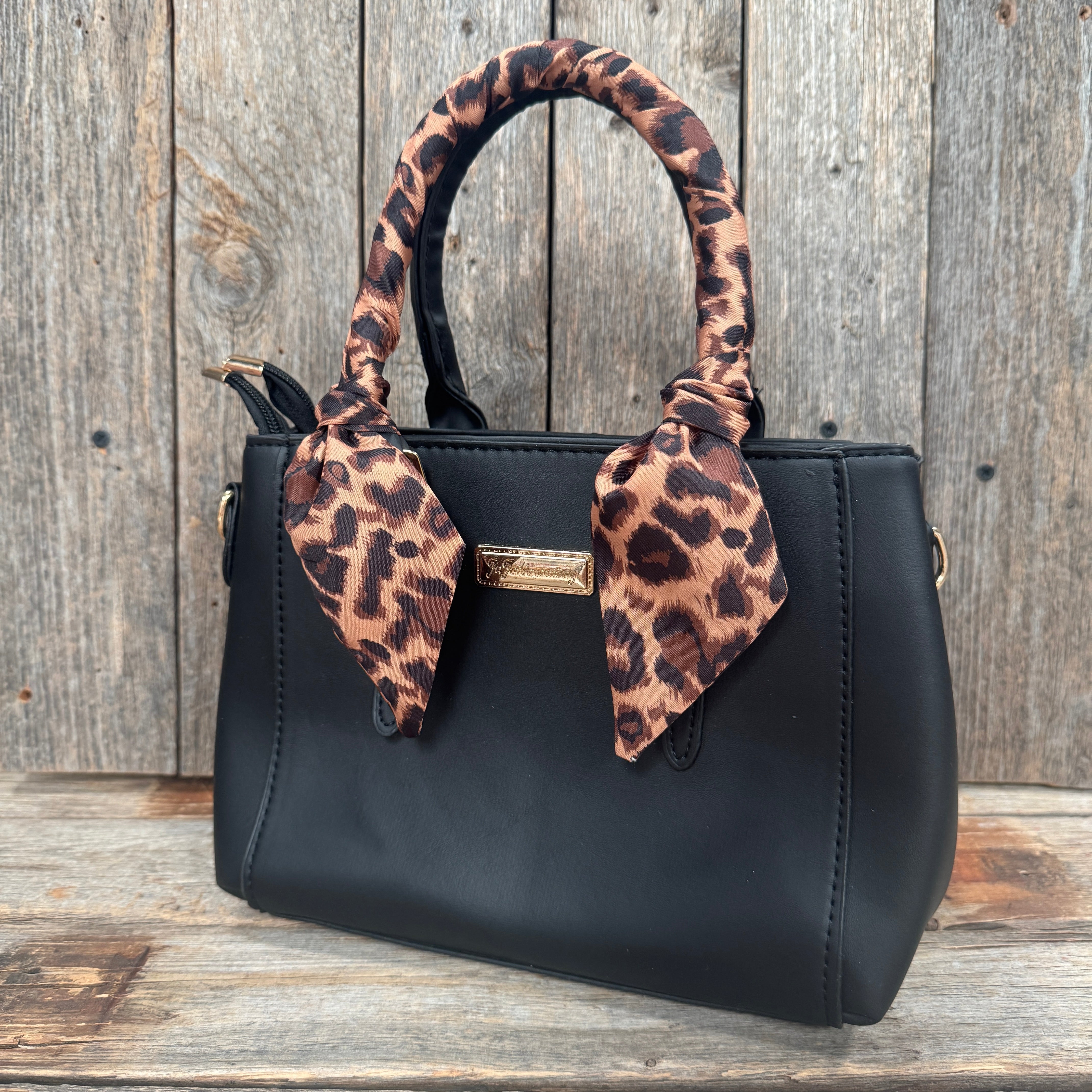A black leather handbag with a leopard print scarf tied around the handle, placed on a wooden surface against a wooden background.