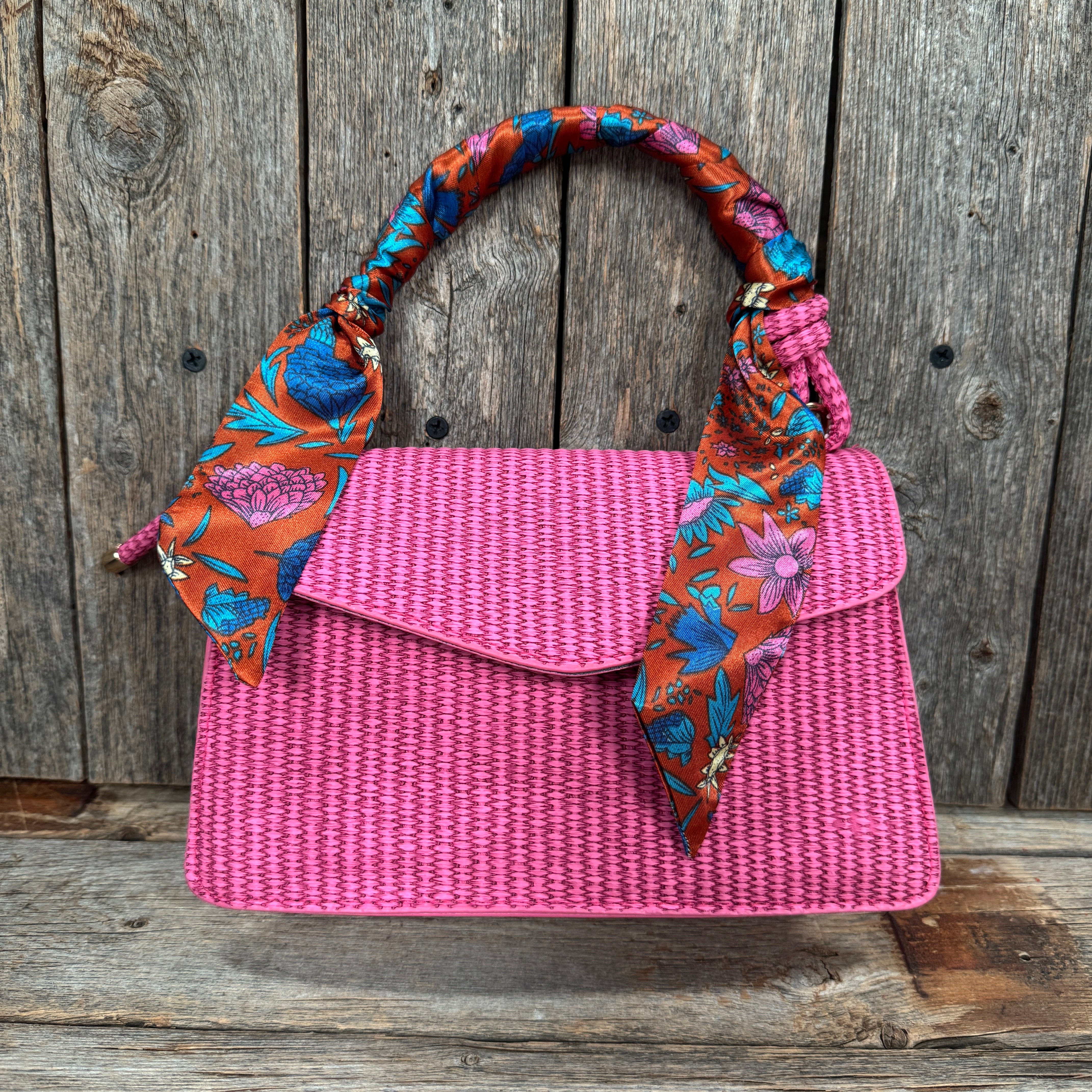 A pink woven handbag with a floral pattern and a colorful scarf tied around the handle, displayed against a wooden background.