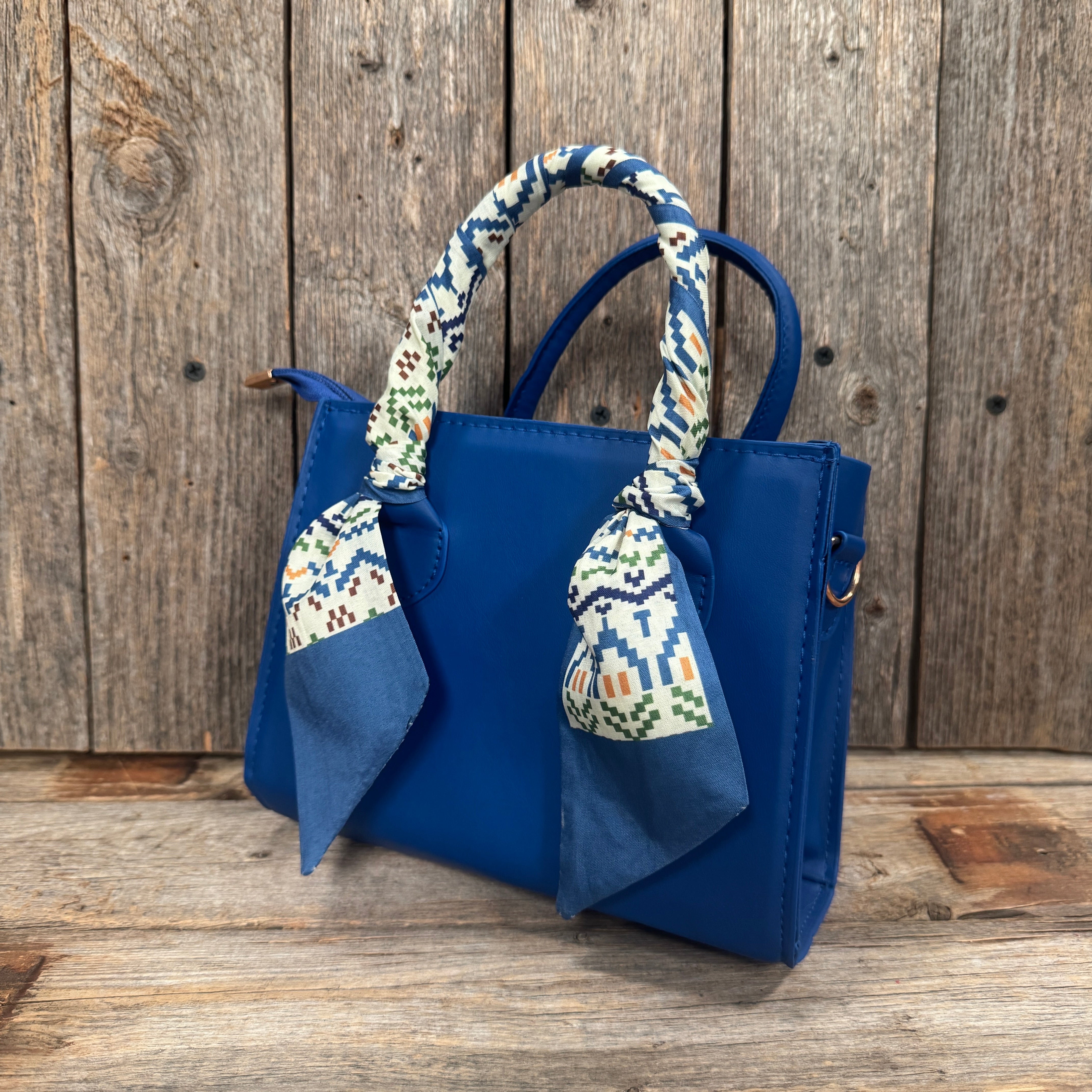 A blue handbag with a patterned scarf tied around the handle, placed on a wooden surface against a wooden background.