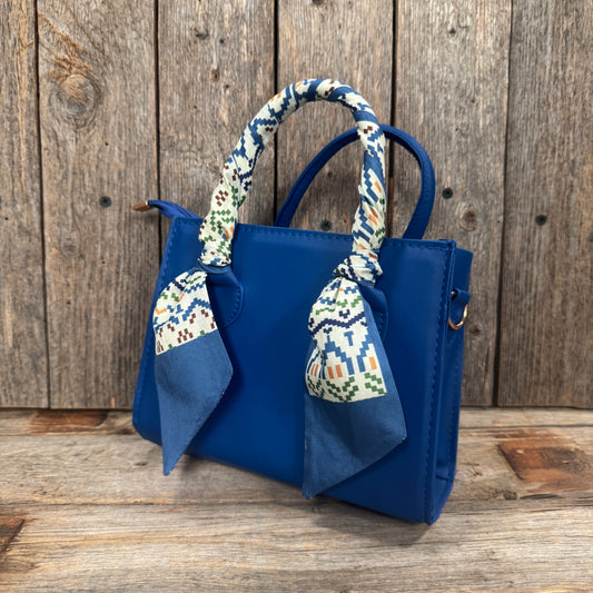 A blue handbag with a patterned scarf tied around the handle, placed on a wooden surface against a wooden background.