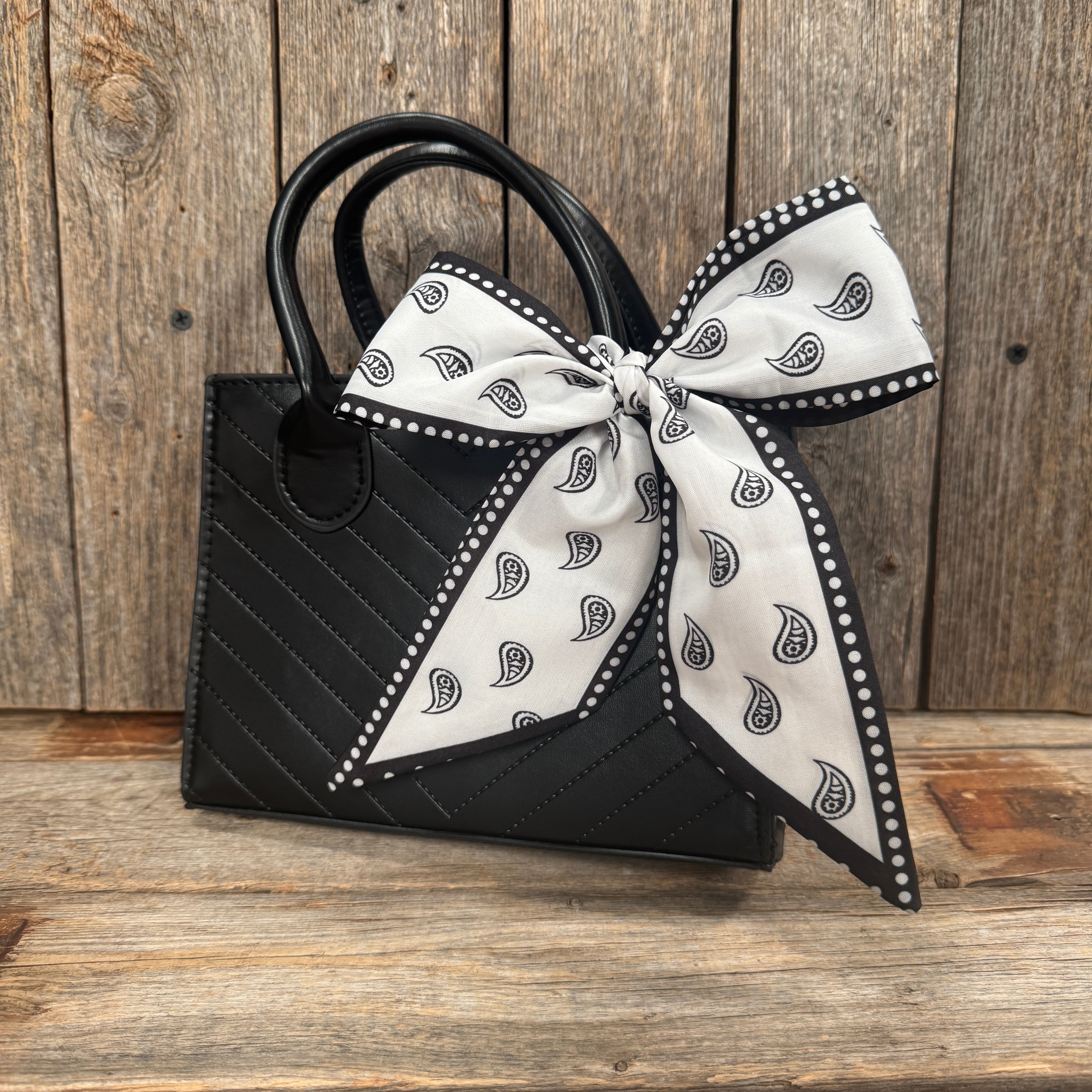 A black quilted handbag with a white paisley-patterned bow tied around the handle, placed on a wooden surface.