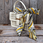 A white handbag with a gold clasp and a colorful patterned scarf tied around it, placed on a wooden surface.