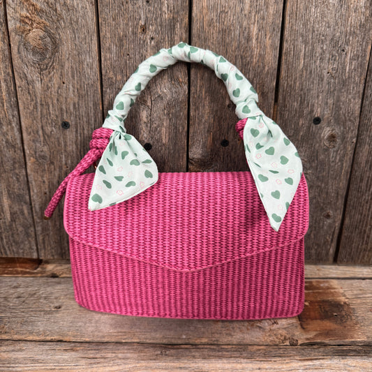 A pink woven handbag with a green and white patterned scarf tied around the handle, placed against a wooden background.