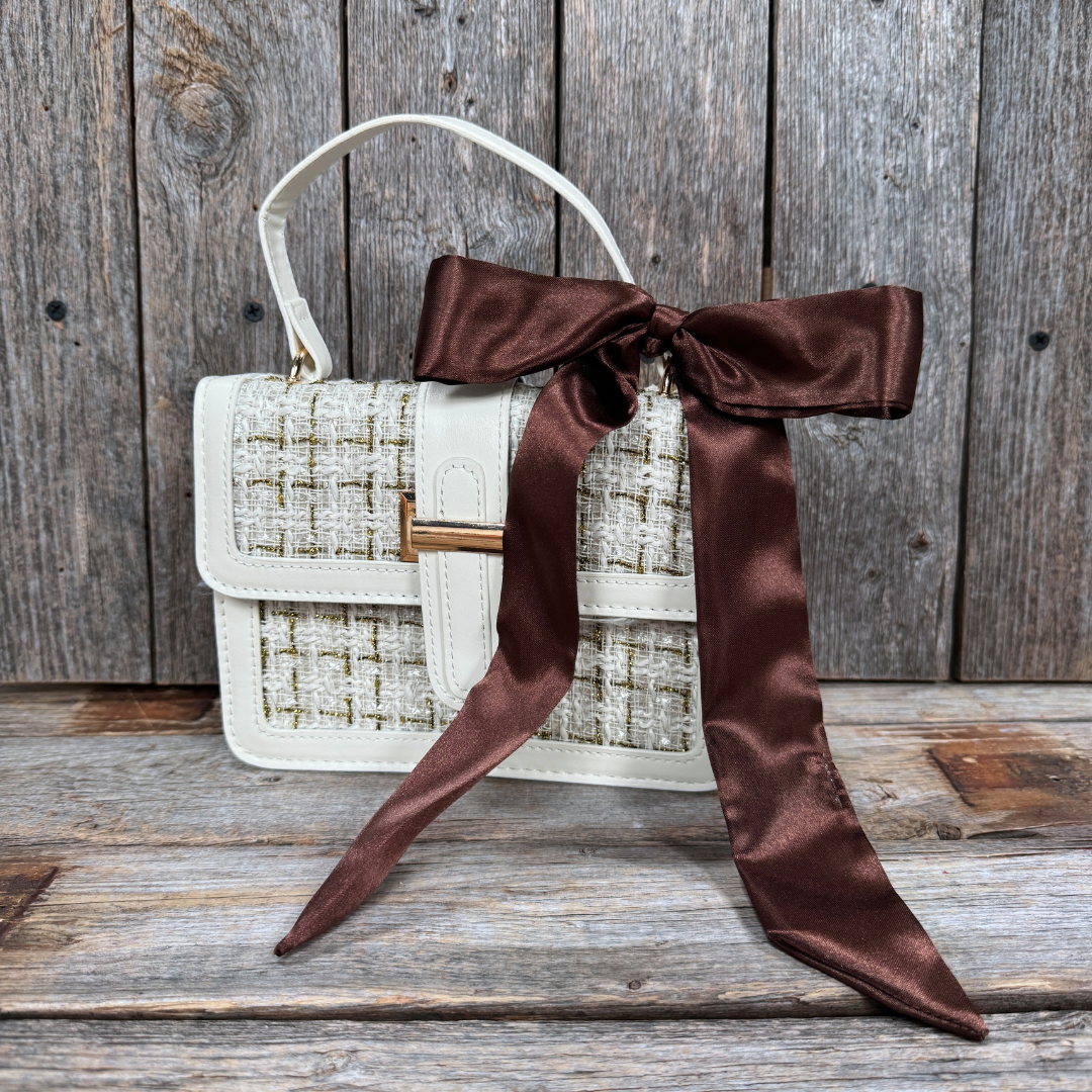 A white handbag with a brown satin bow tied around it, placed on a wooden surface.
