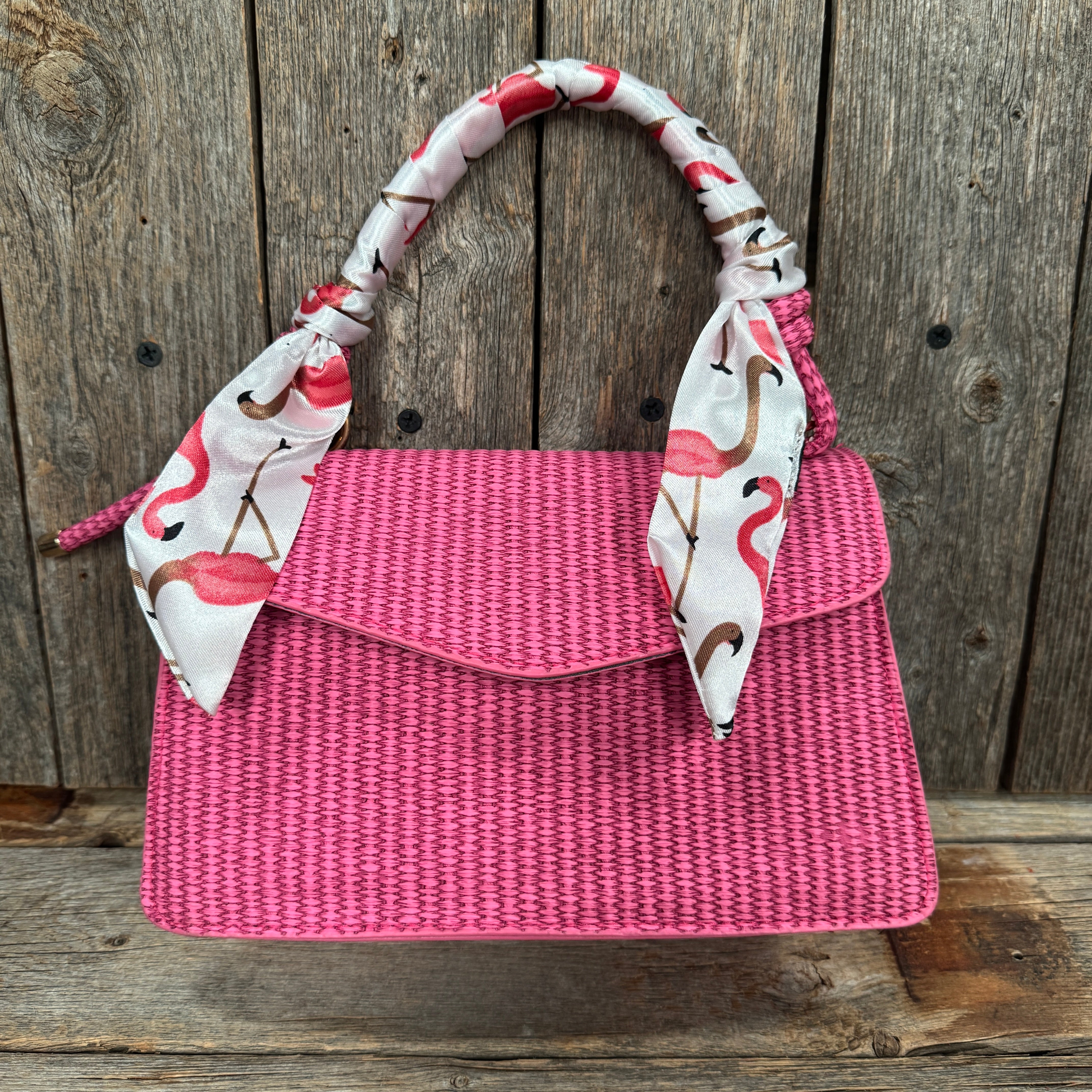 A pink woven handbag with a white and pink flamingo patterned scarf tied around the handle, displayed against a wooden background.