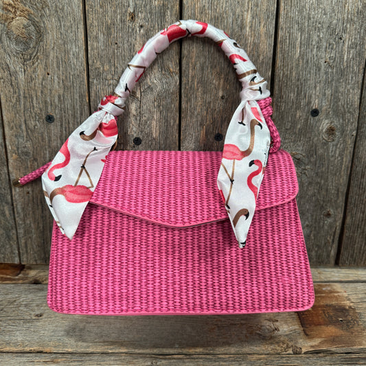 A pink woven handbag with a white and pink flamingo patterned scarf tied around the handle, displayed against a wooden background.