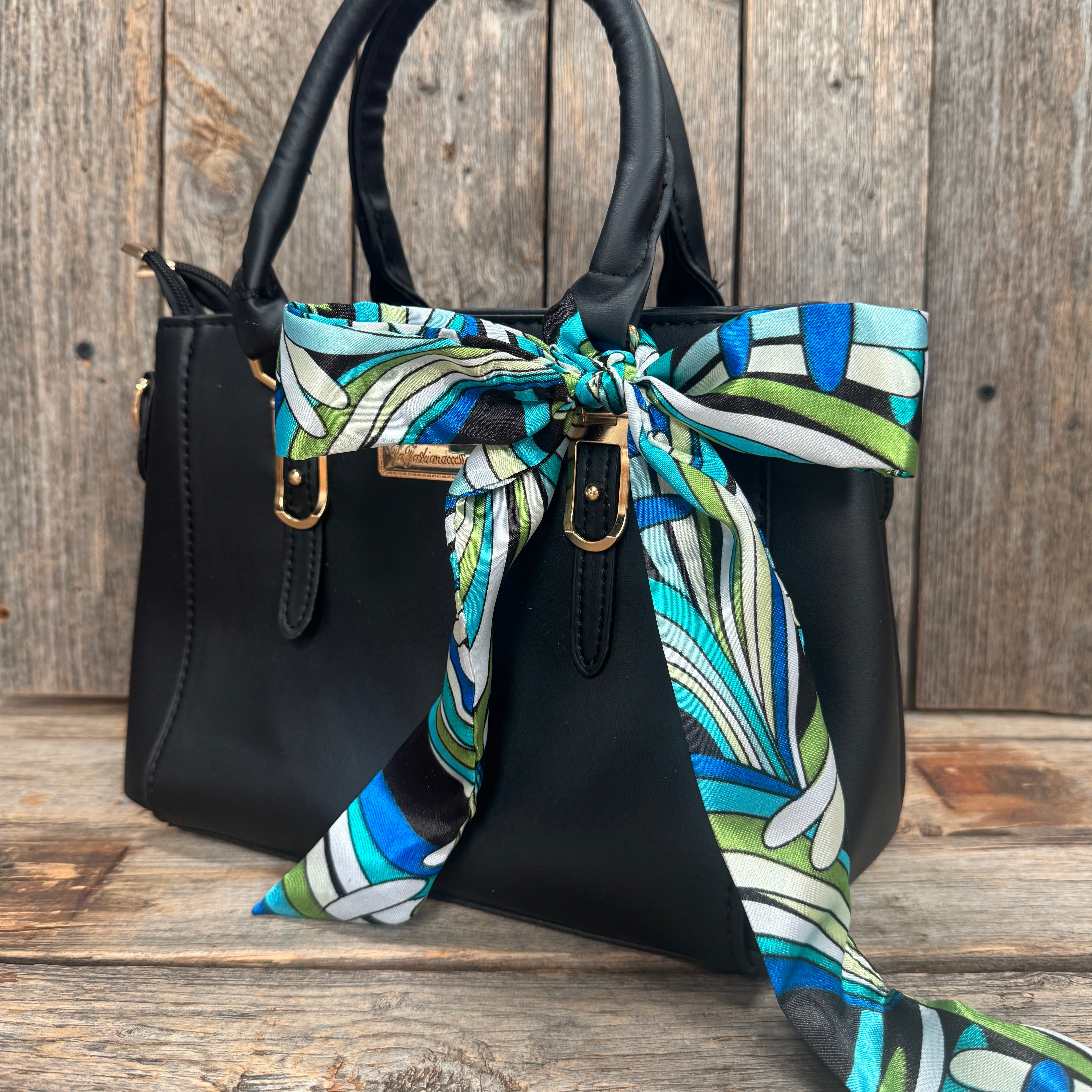 A black handbag with a colorful patterned scarf tied around the handle, placed on a wooden surface.