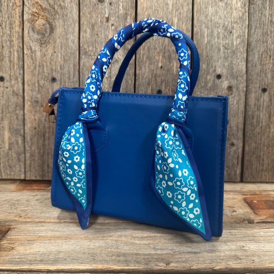 A blue handbag with a floral patterned scarf tied around the handles, placed on a wooden surface.
