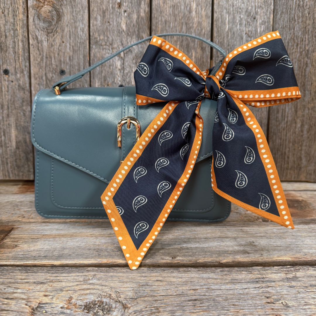 A blue handbag with a black and orange paisley-patterned scarf tied around the handle, placed on a wooden surface.
