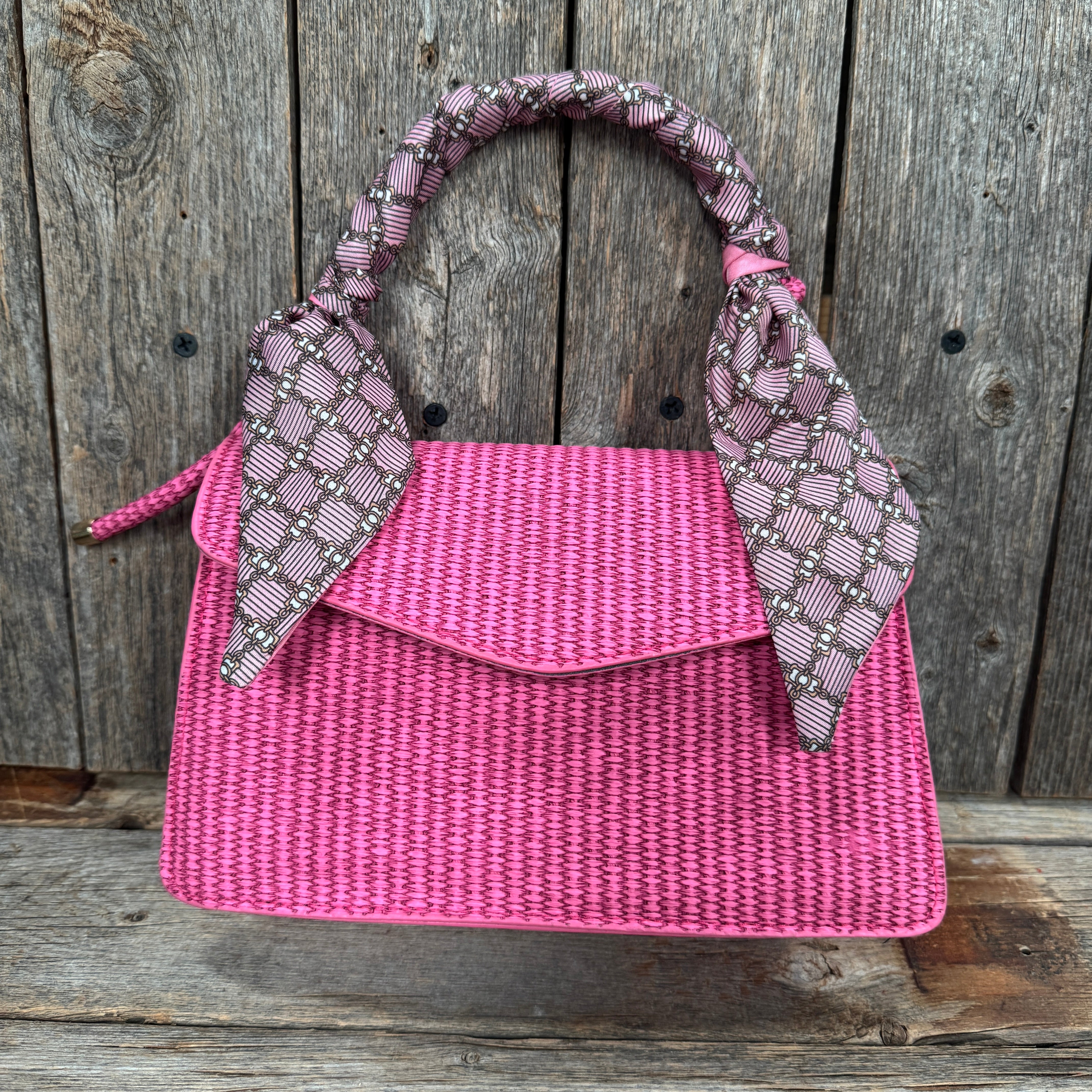 A pink woven handbag with a patterned scarf tied around the handle, displayed against a wooden background.
