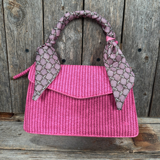 A pink woven handbag with a patterned scarf tied around the handle, displayed against a wooden background.