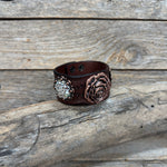A dark brown leather cuff bracelet featuring a textured rose motif and a rhinestone-embellished floral accent, displayed on a rustic weathered wood background.