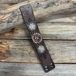 A dark brown leather bracelet featuring a central rose design, two floral medallions with rhinestones, and silver-tone metal studs, displayed on a rustic wooden background.