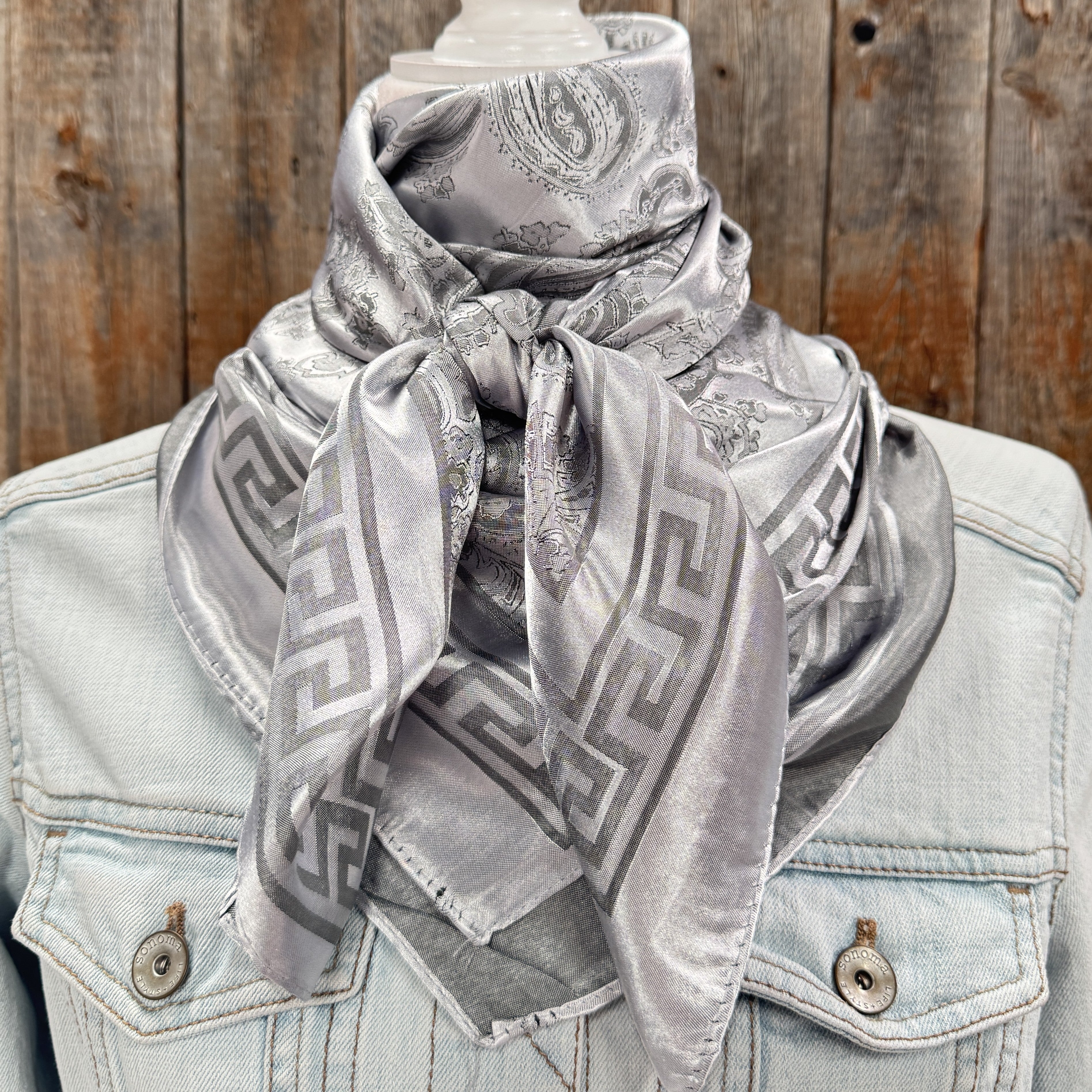 A silver floral jacquard pattern scarf displayed on a mannequin wearing a denim jacket.