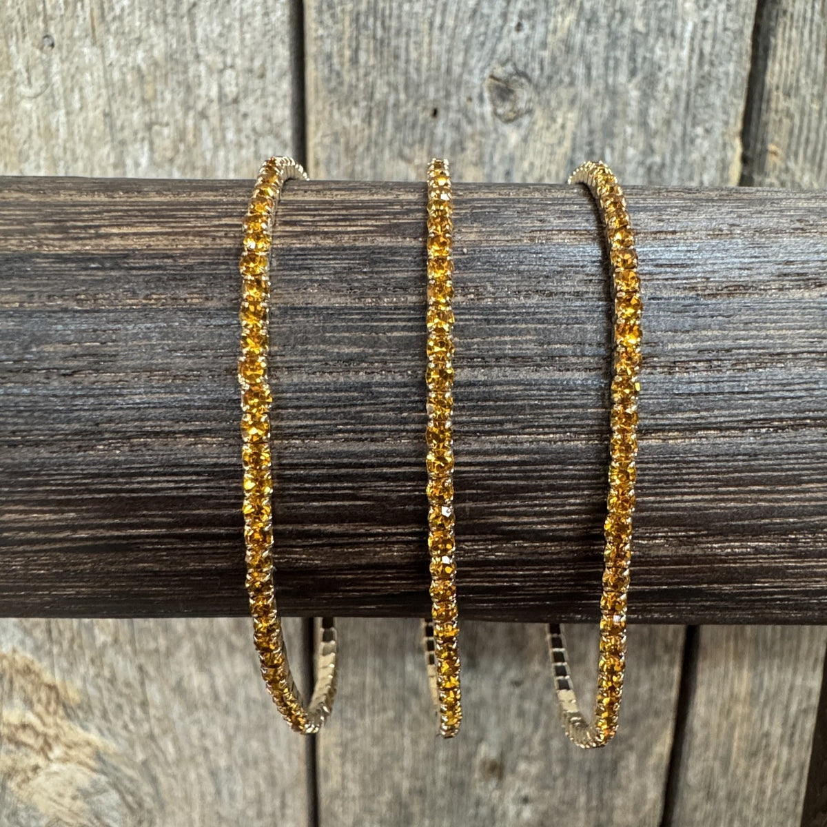 Three gold-toned bangle bracelets covered in small clear stones, displayed on a dark wooden dowel against a rustic wood plank background.