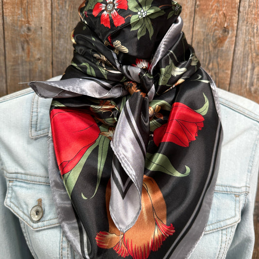 A western wild rag scarf with silver, red, and green floral patterns tied around the neck, displayed on a person wearing a denim jacket.