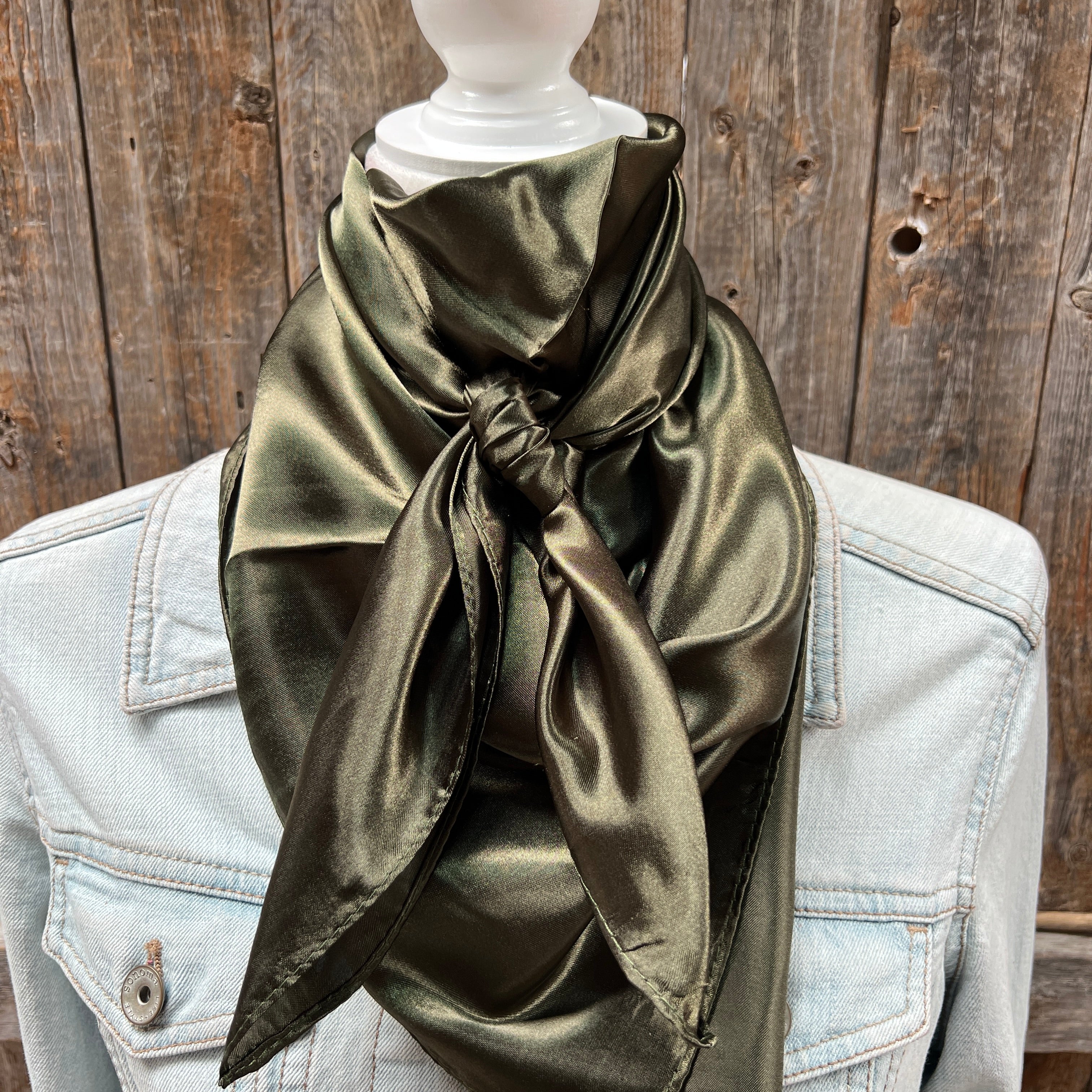 A solid olive green wild rag scarf displayed on a mannequin wearing a denim jacket, against a wooden backdrop.