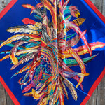Colorful feather pattern wild rag scarf 
on a blue background with red border, on a wooden surface