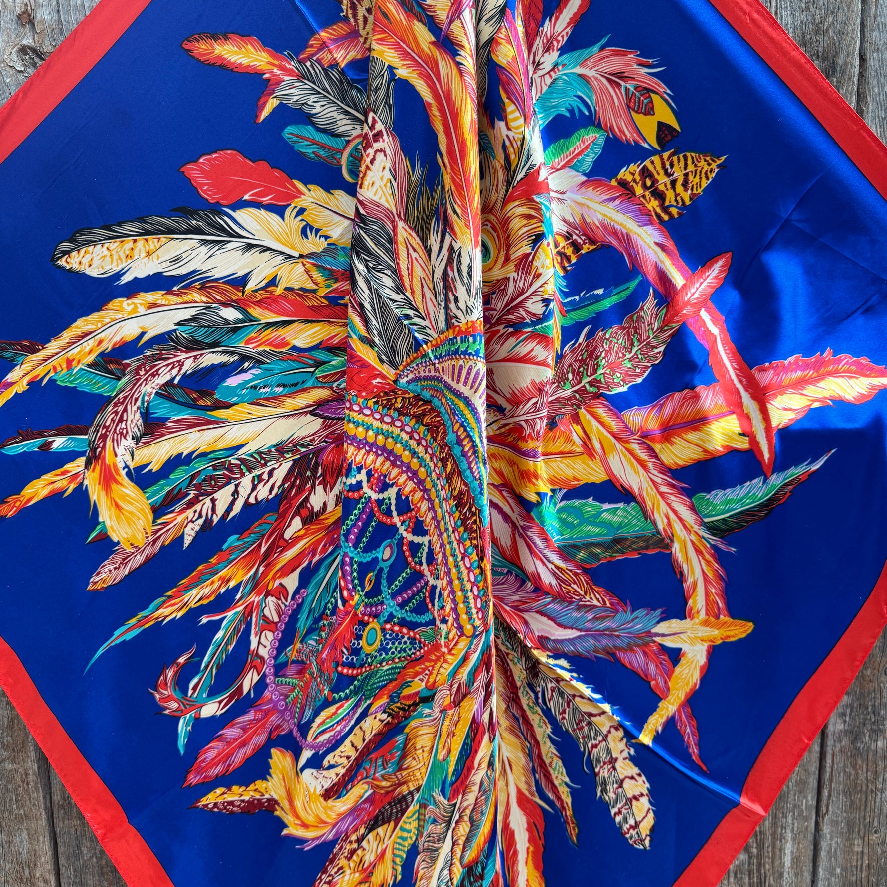 Colorful feather pattern wild rag scarf 
on a blue background with red border, on a wooden surface