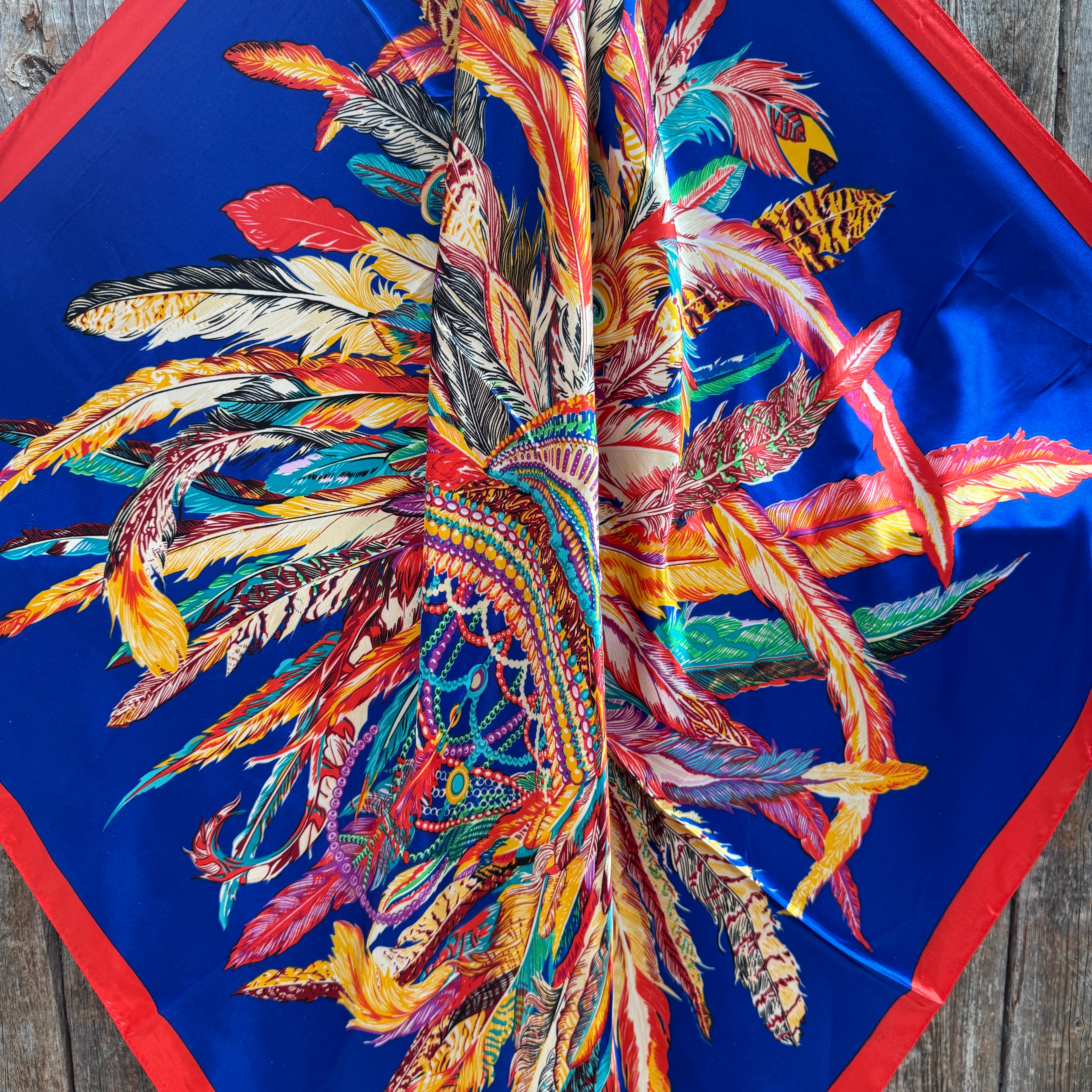 Colorful feather pattern wild rag scarf 
on a blue background with red border, on a wooden surface