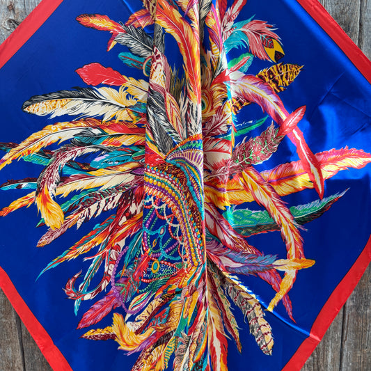 Colorful feather pattern wild rag scarf 
on a blue background with red border, on a wooden surface
