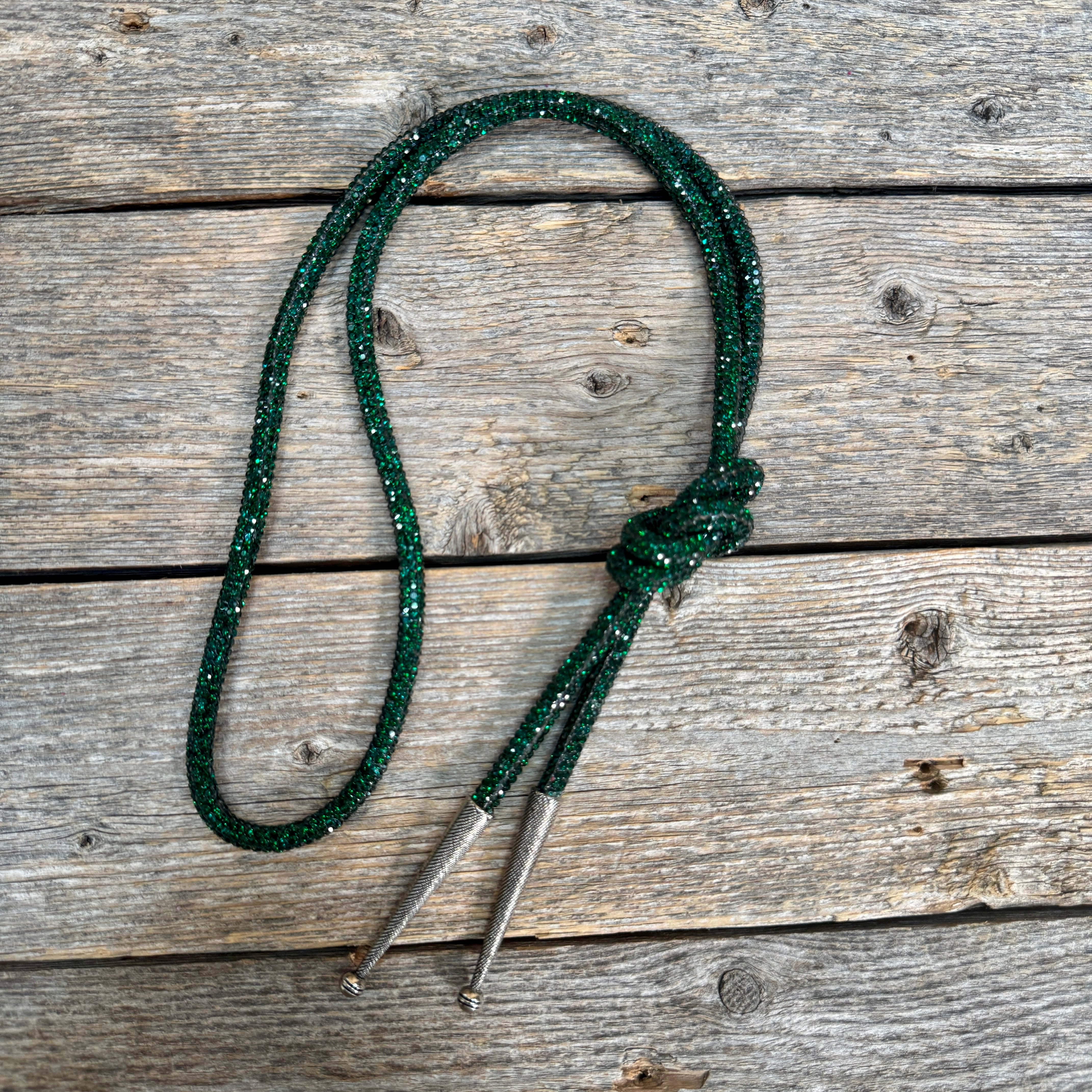 A dark green glitter rope bolo tie with silver-toned metal tips, laid flat on a rustic weathered wood plank background.