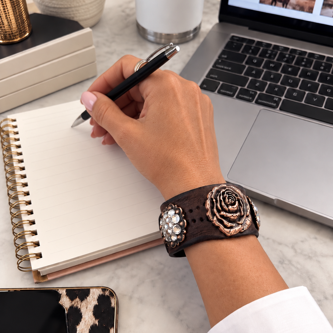 A close-up lifestyle shot of a person's hand writing in a spiral notebook on a marble desk, wearing a brown leather rose bracelet with rose gold and crystal embellishments.