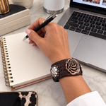 A close-up lifestyle shot of a person's hand writing in a spiral notebook on a marble desk, wearing a brown leather rose bracelet with rose gold and crystal embellishments.