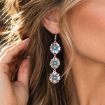 A close-up lifestyle shot of a woman wearing a long, multi-stone dangle earring. The earring features silver-toned metal settings with light blue and white stones.