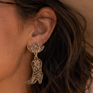 A close-up lifestyle shot of a woman wearing a tan cowboy hat, black aviator sunglasses, and a dangling gold cowboy boot earring with intricate cutout detailing.