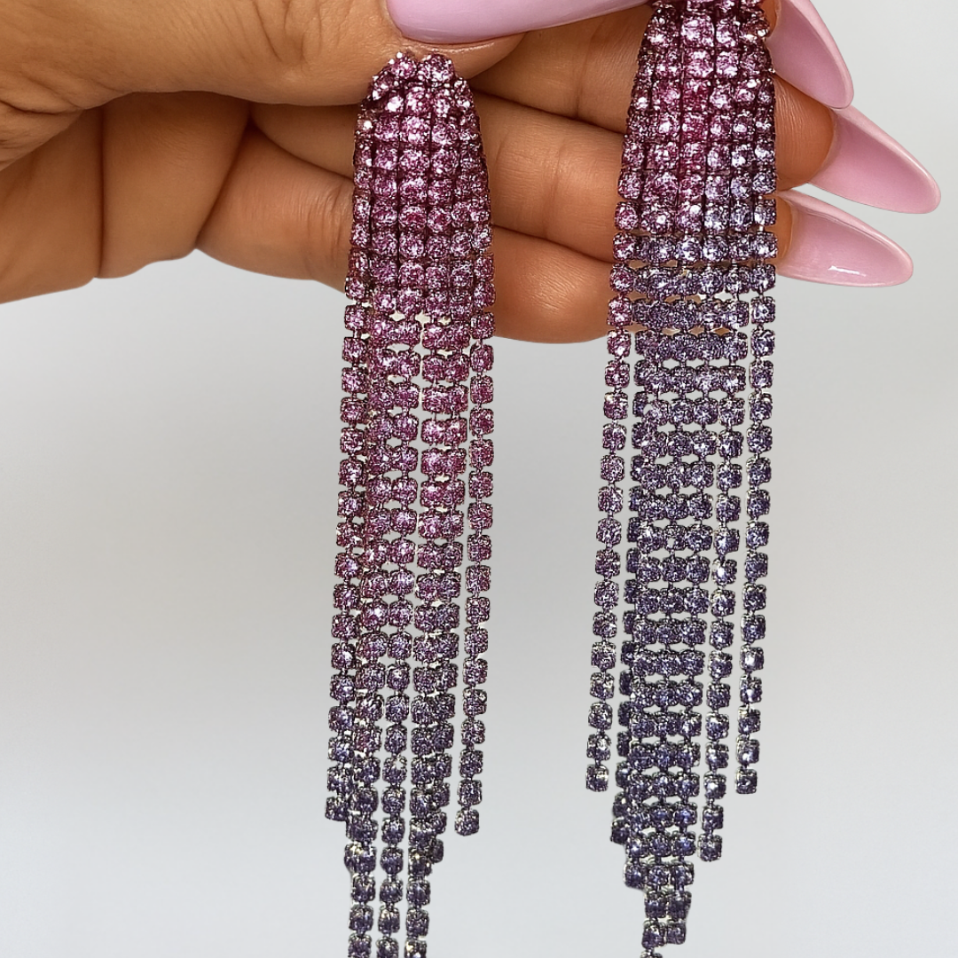 Long, dangling earrings with pink gemstones held by a hand against a white background