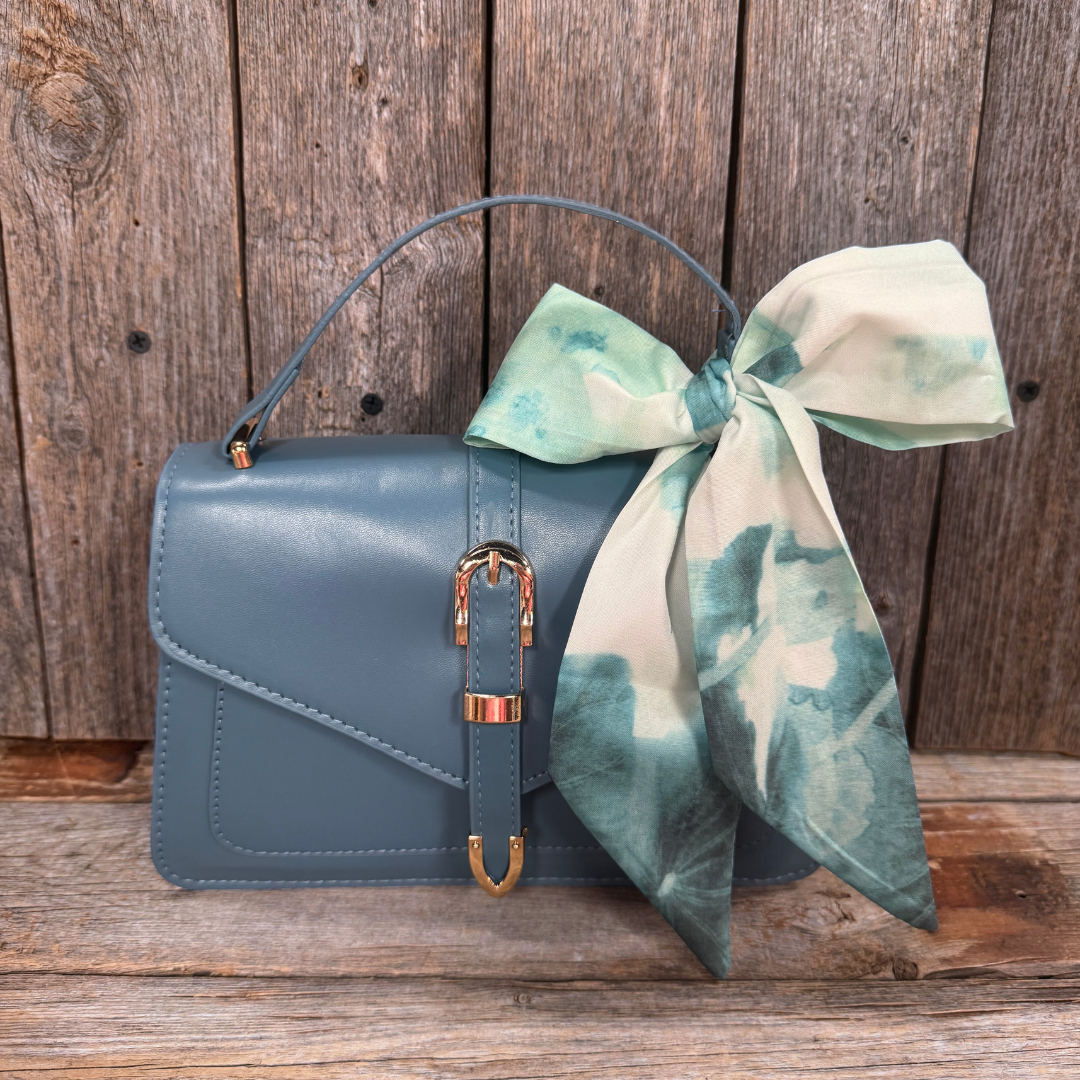 A blue handbag with a gold buckle and a green and white patterned scarf tied around the handle, placed on a wooden surface.