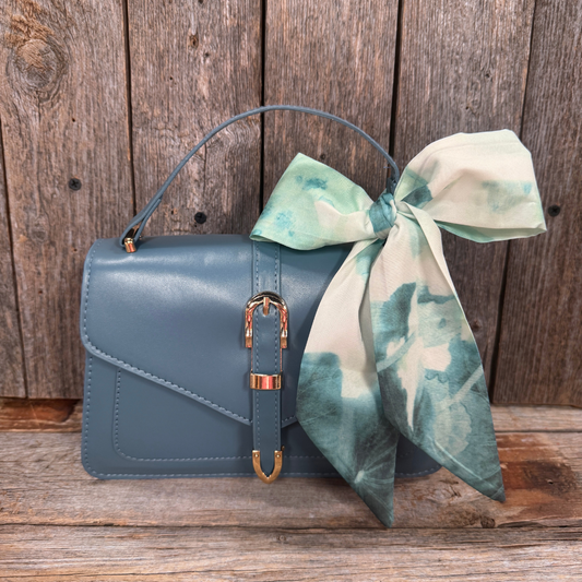 A blue handbag with a gold buckle and a green and white patterned scarf tied around the handle, placed on a wooden surface.