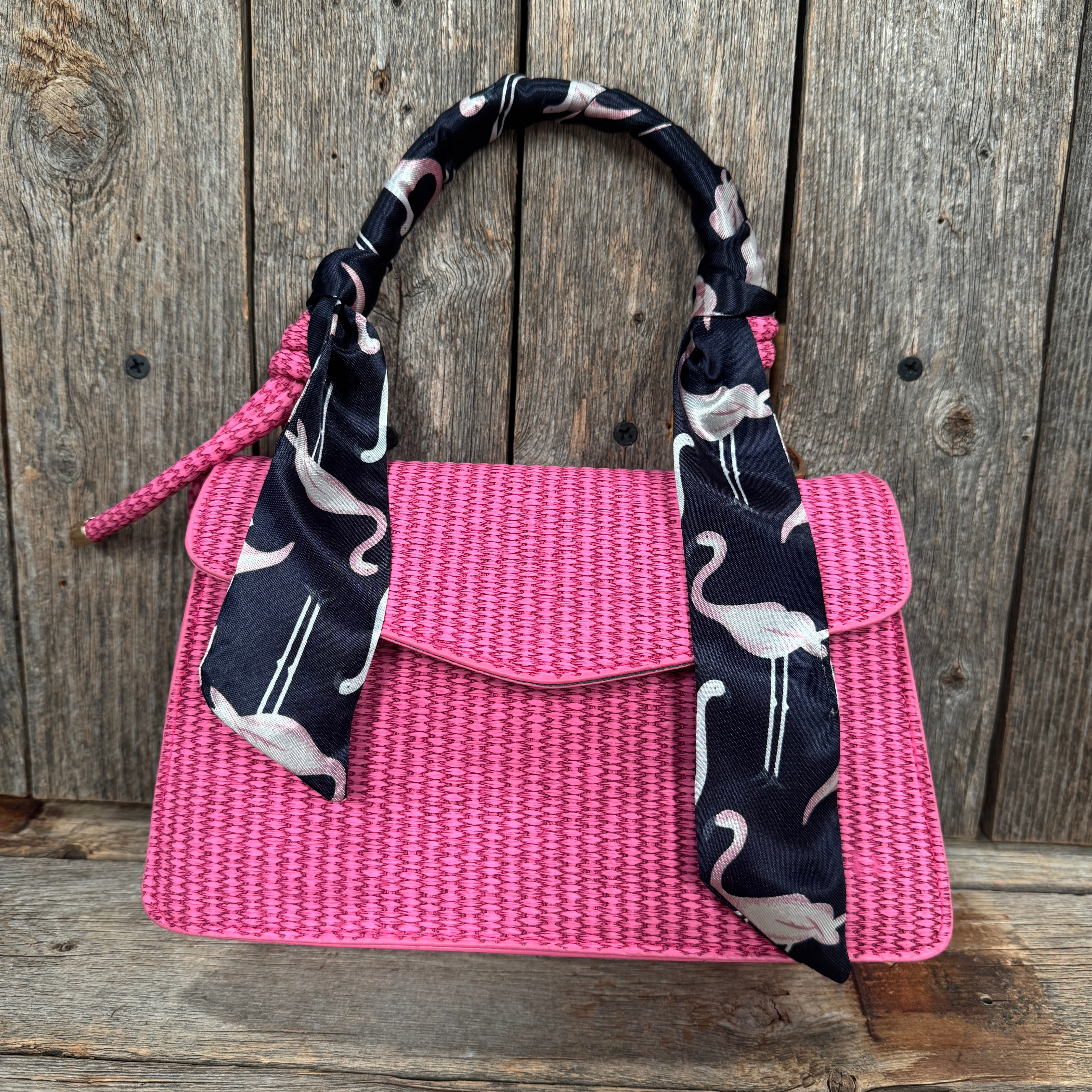A pink woven handbag with a black and white patterned scarf tied around the handle, placed against a wooden background.
