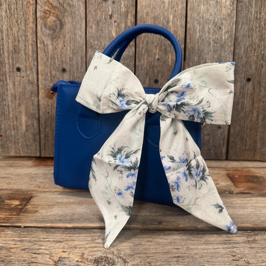 A blue handbag with a white floral patterned ribbon tied around it, placed on a wooden surface.