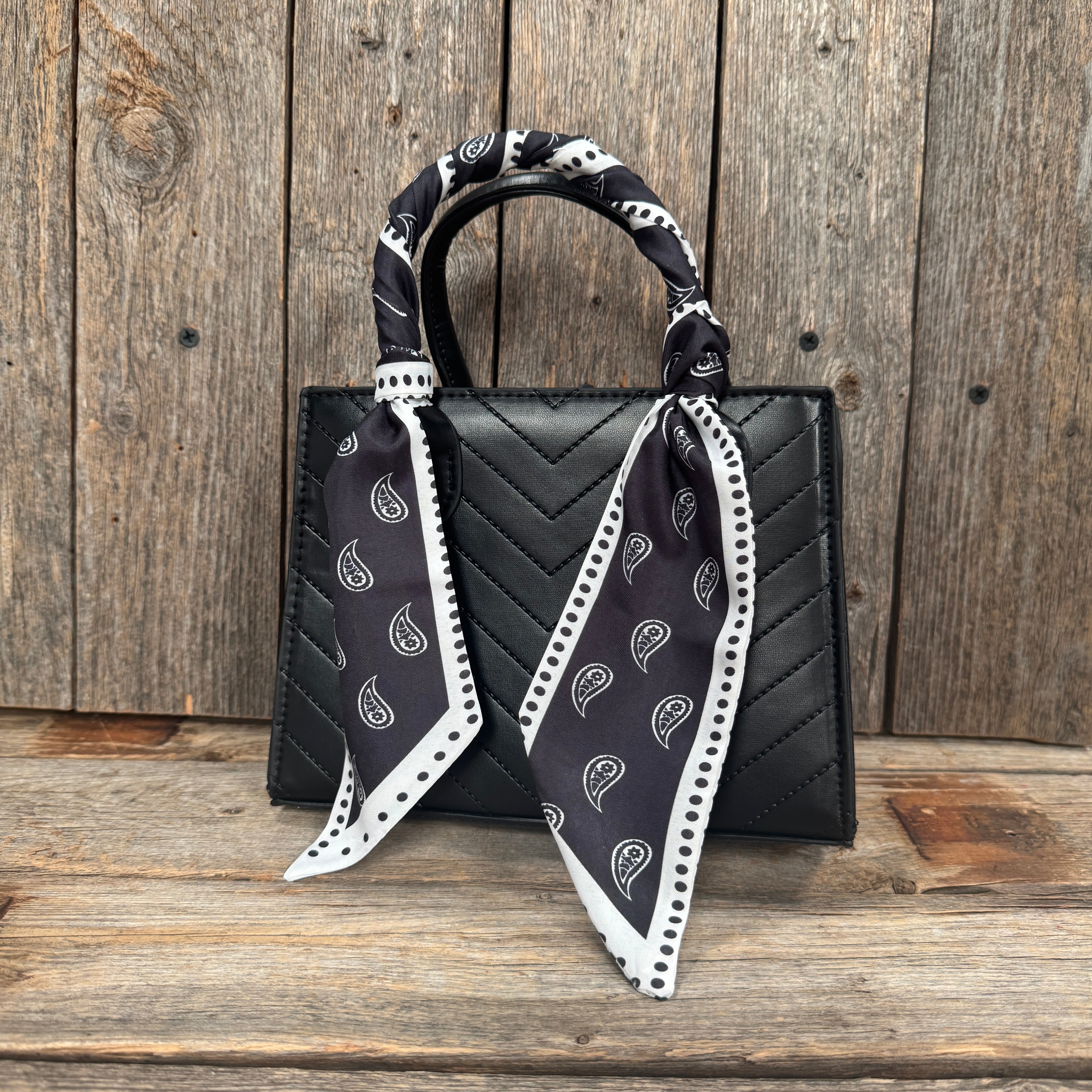 A black handbag with a black and white patterned scarf tied around the handle, placed on a wooden surface against a wooden background.