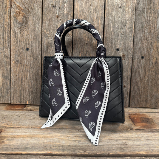 A black handbag with a black and white patterned scarf tied around the handle, placed on a wooden surface against a wooden background.