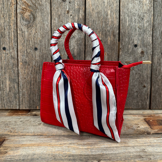 A red handbag with a red and white striped ribbon tied around the handle, placed on a wooden surface against a wooden background.